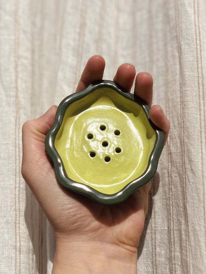 wavy rimmed soap dish held in the palm of a hand against a fabric background. the soap dish is dark green on the outside with a lime green inside