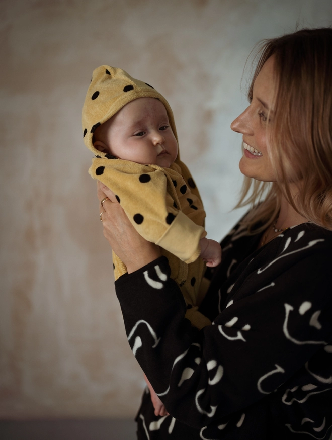 Baby wearing the Another Fox Camel Terry Towel Spot Baby Sleepsuit held by a woman.