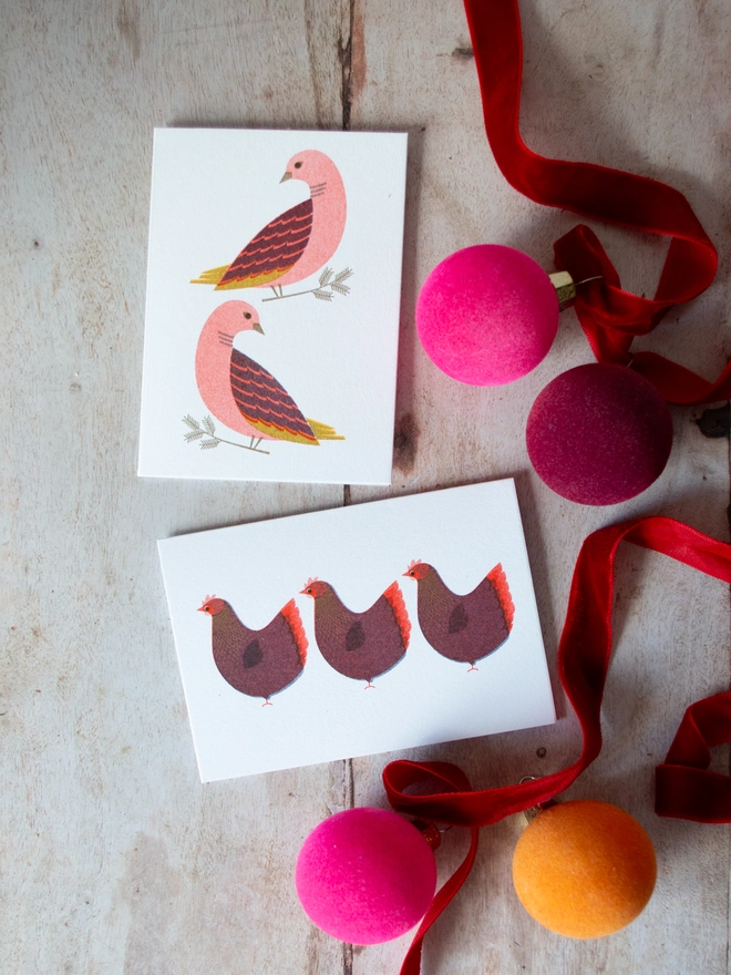 Two lavish and luxury feeling risograph printed mini Christmas cards featuring the traditional folklore characters, two turtle doves and three French hens are placed on a wooden table with jewel toned festive Christmas baubles and ribbons surrounding it to give a maximalist festive feel.
