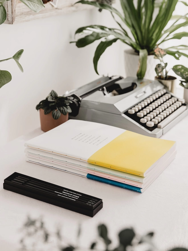Stack of hand stitched notebooks on a desk in yellow, pale green, pale pink, blue and white on a desk with a typewriter and pencil set