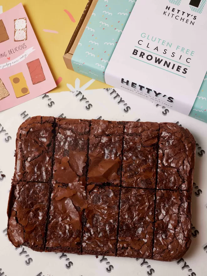 Ten slices of gluten free classic fudge brownies in a flat lay with branded box and postcard