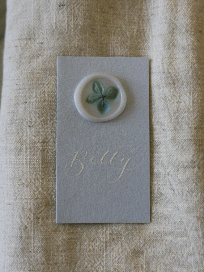 Pale blue place card with white calligraphy and vellum white wax seal with blue hydrangea petals on linen napkin 