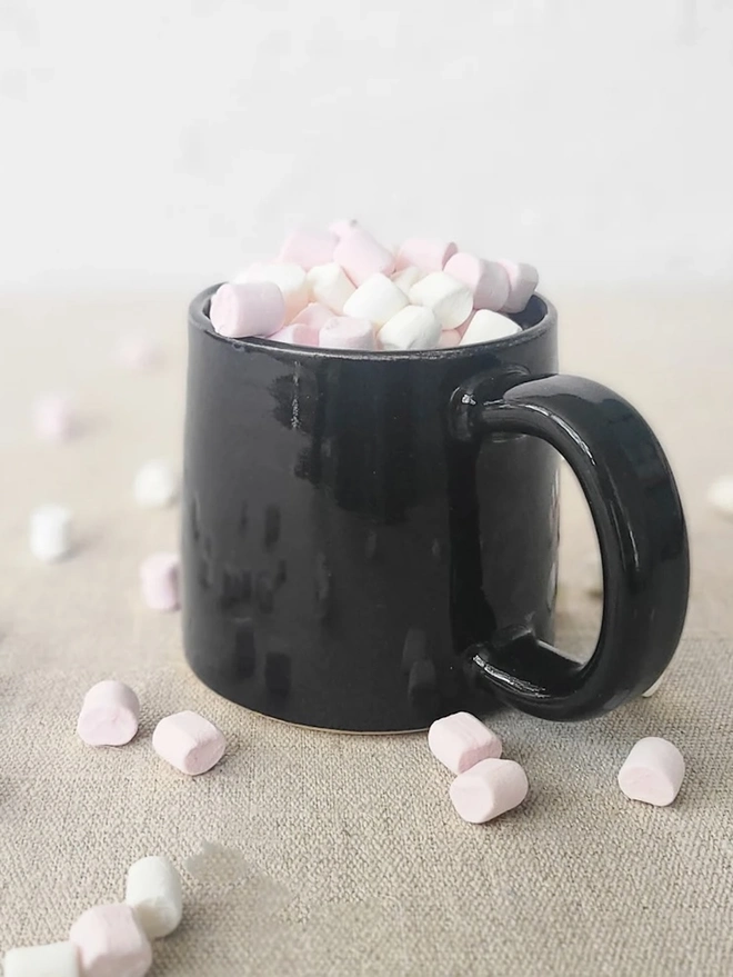 Ceramic Standard Mug, a colourful ceramic mug sitting on a plain surface against a grey backdrop. It is accompanied by marshmallows. 