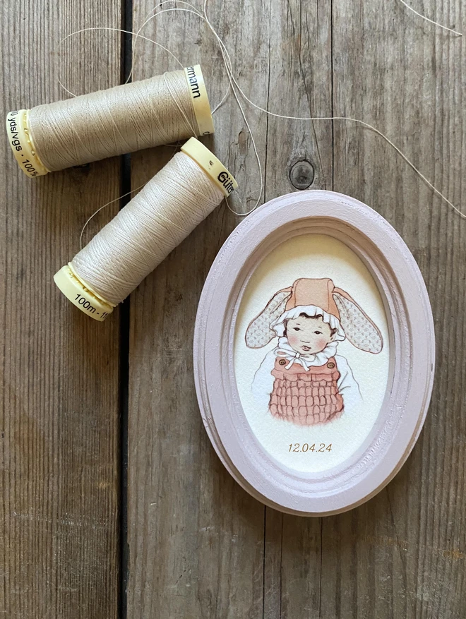 a photograph of a miniature hand drawn personalised easter portrait featuring a vintage style hand drawn story book style illustration of a little Chinese asian Japanese Korean baby girl wearing a bunny eater bonnet framed in a handmade oval dusty pink wooden frame perfect for easter