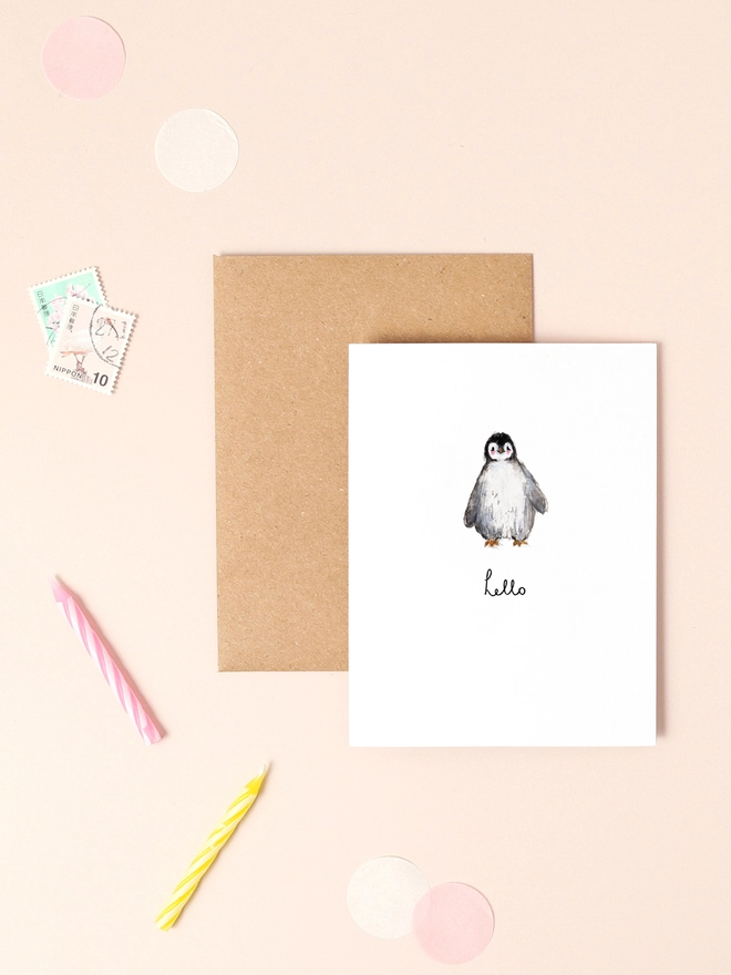 A cute illustration of a small penguin standing upright with a cheerful expression, holding a tiny sign that reads "Hello." The penguin's black and white feathers and orange beak are depicted in soft, charming detail. The design has a light, minimalist style with a pastel background, perfect for sending a sweet and simple greeting