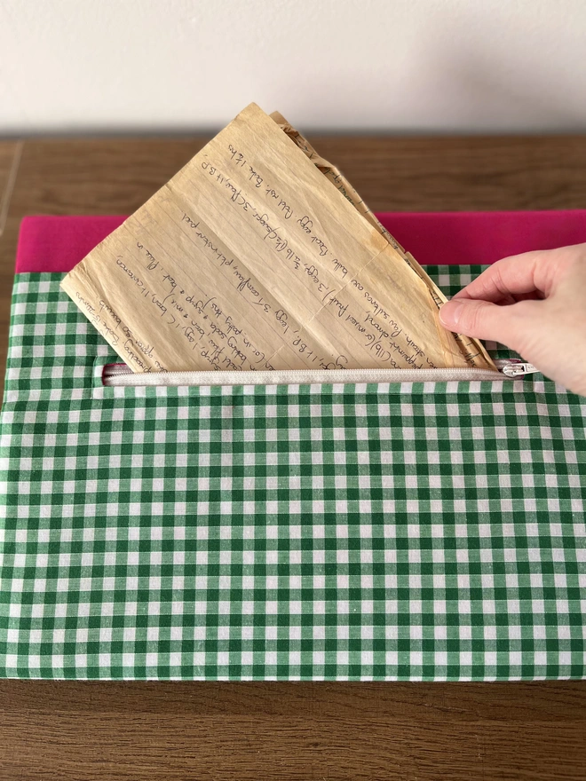Close-up of hand putting old recipe notes into open zip pocket on back of green gingham with pink spine fabric cover of recipe binder. The zip is white. and binder is lying flat on wooden bench.