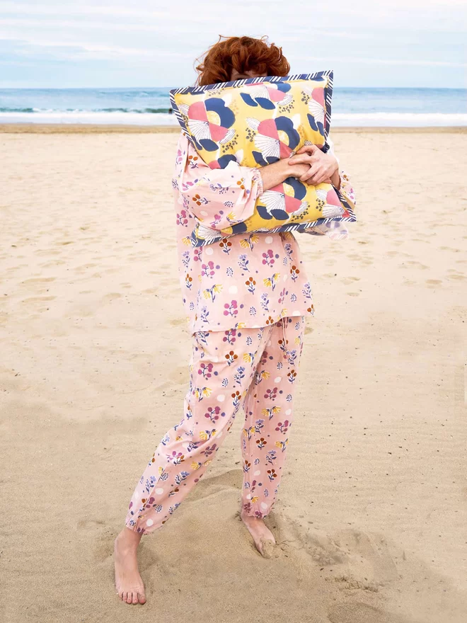 Red haired model wearing a block printed dusty pink floral pyjama and covering her face with a yellow cushion featuring a large scale floral design in pink, white and navy colours