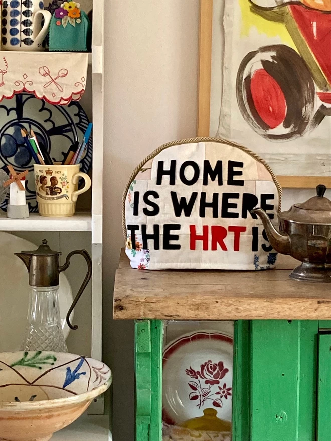 'Home is where the HRT is' tea cosy shown on a dresser, displaying crockery and a tea pot.