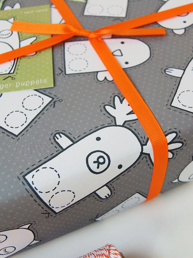 A gift wrapped in grey wrapping paper that is covered in drawings of animal finger puppets that can be cut out and coloured lays on a white desk.