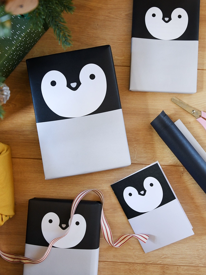Several penguin greetings cards and gifts wrapped in penguin wrapping paper lay on a wooden floor.
