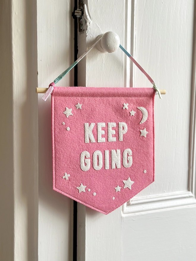 A pink felt banner with the words Keep Going appliquéd on with white felt letters, hangs from a wooden dowel and cotton ribbon on a white door handle.