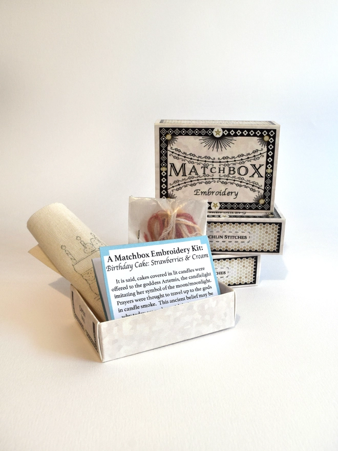 The kit supplied in a beautifully designed matchbox, pre-printed fabric and small instruction leaflet neatly folded to sit inside, along with the yarns and needle housed in a small glassine packet.  A stack of matchboxes sit in the background.