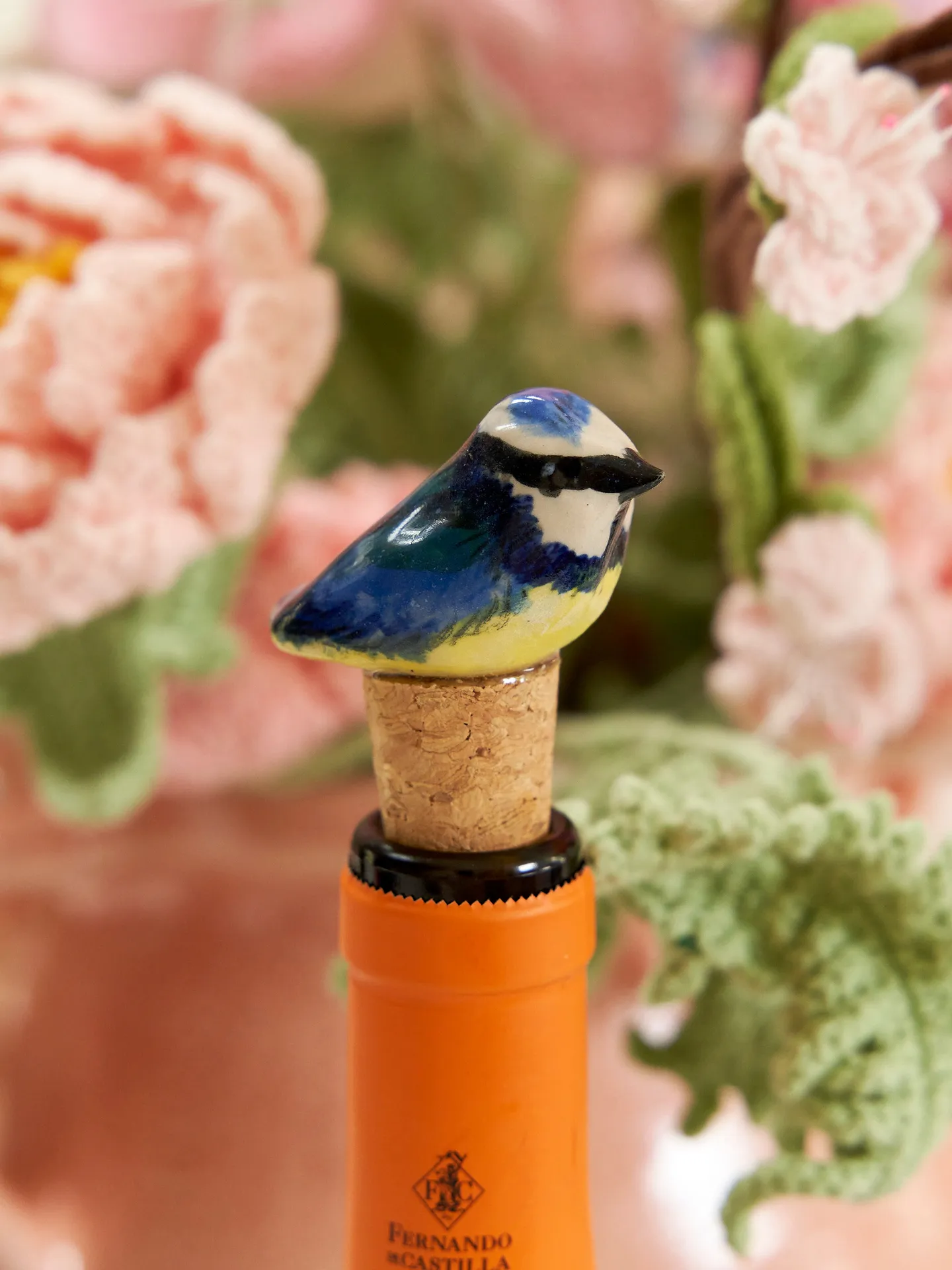 Blue Heart Ceramic Bird Bottle Stop, a bird bottle stop sitting on a glass bottle surrounded by floral decorations. 