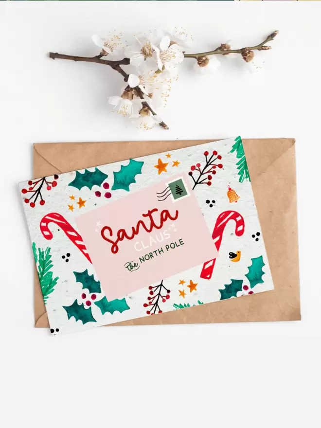 In the centre of the image is a card with a postcard addressed to Santa Clause surrounded by holly, star and candy cane illustrations. Above the card is a white flower sprig.