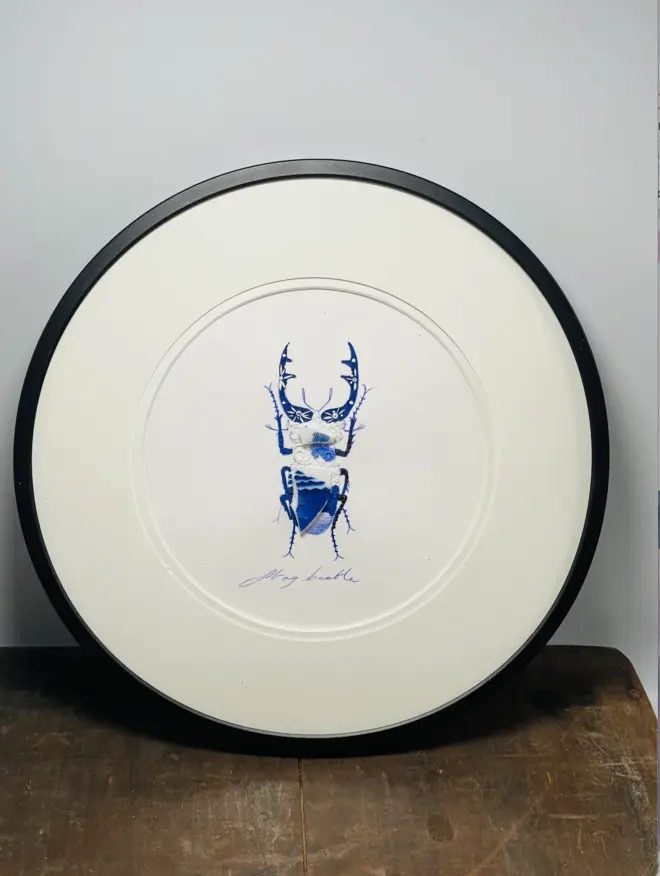 Hand-drawn, painted, mosaic clay, blue Willow Tree pottery insect on white double mounted card, in a black circular frame.