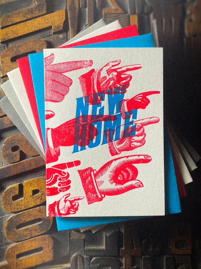 A double-sided, hand-printed letterpress artisan moving house announcement card with bold blue “New Home” typography and fluorescent red pointing hand illustrations (fists) on thick premium white card.