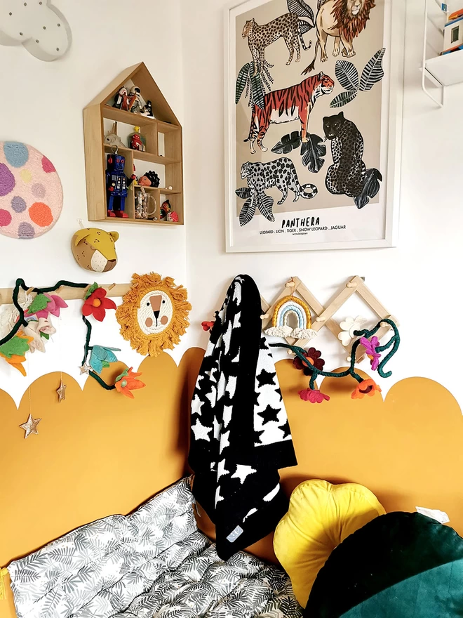 A modern childrens room with a cosy cushioned reading corner and half scalloped ochre walls. The walls are adorned with fun childrens decor, and on a wall peg hangs a black and white knitted star baby blanket.