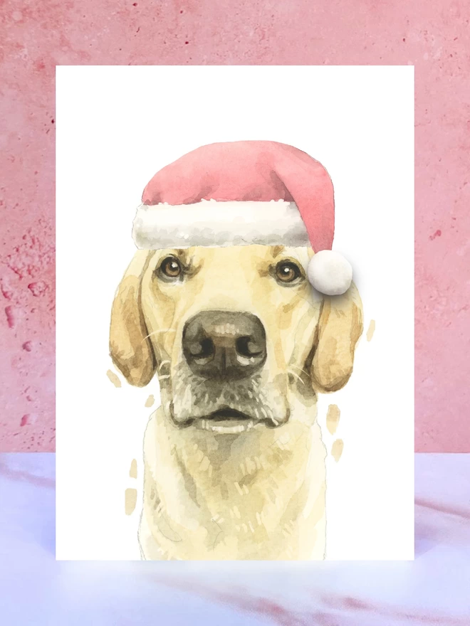 A Christmas card featuring a hand painted design of a yellow labrador, stood upright on a marble surface. 