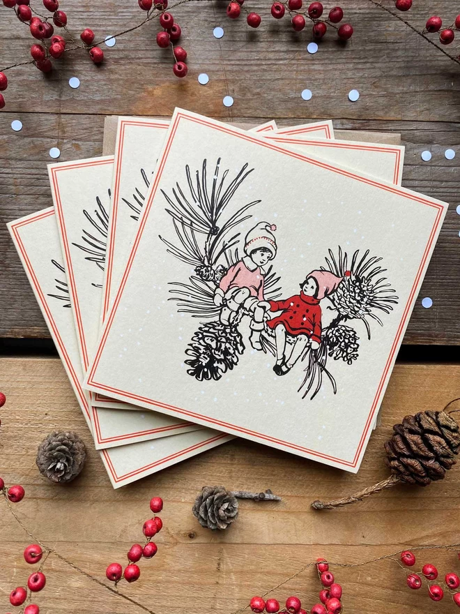 4 Christmas cards showing an ink drawing in a vintage children's book style of winter pixies sat on a pine cone branch