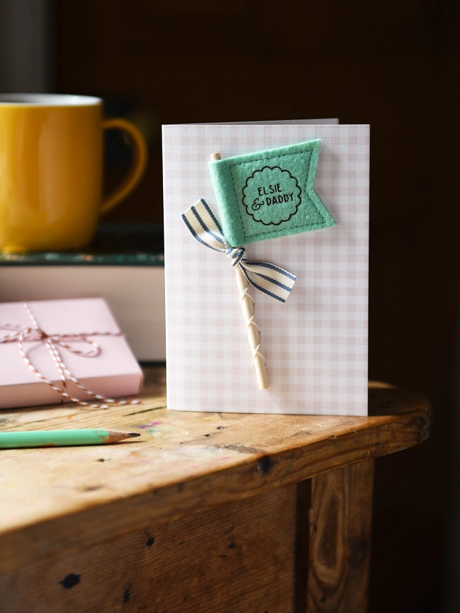 Personalised Father’s Day card with mustard felt flag reading “Henry & Daddy”, handmade keepsake card with ribbon and wooden stick detail. The card stands on a wooden table beside a book, mug, pencil, and wrapped gift.
