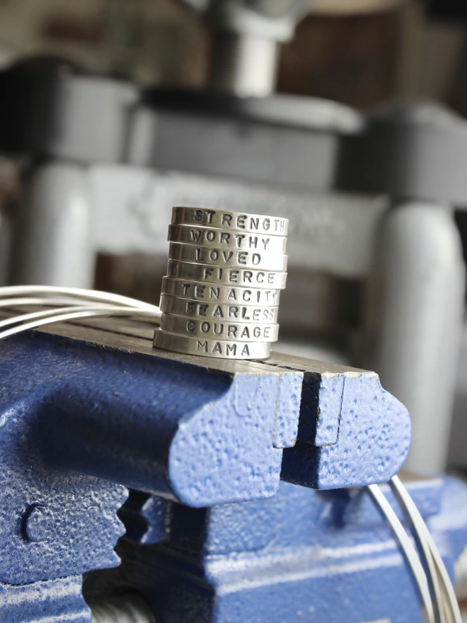 A stack of sterling silver plain band rings, each stamped with a single word, arranged in a tower and resting on a blue bench vice with a coil of wire and a rolling mill in the background.