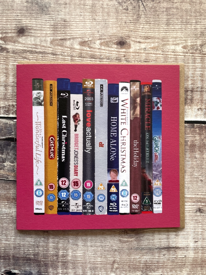 Christmas card design of stacked festive movie spines (Elf, Home Alone, Love Actually, The Holiday, Gremlins) on a wood backdrop.