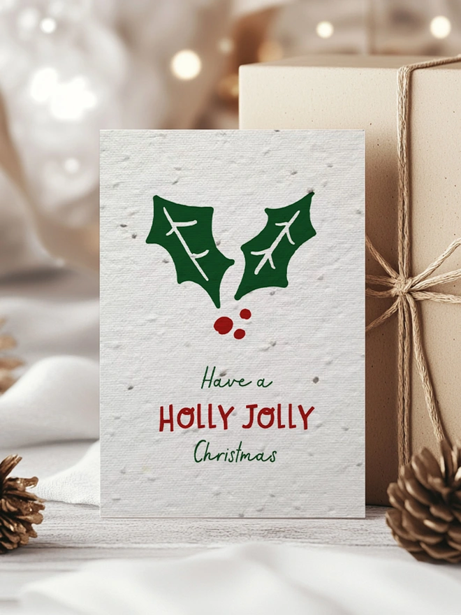 Christmas card with a holly leaf on with 'Have a Holly Jolly Christmas'. Behind the card is some presents, a pine cone and pine leaves.