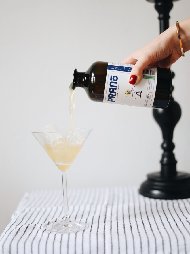 Hand pouring a pale orange drink from a dark brown bottle into a martini glass, sitting atop a striped tablecloth