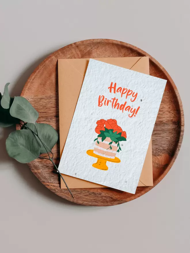 Plantable Card with Happy birthday and an illustration of a cake with roses on, on a wooden tray next to a Eucalyptus branch