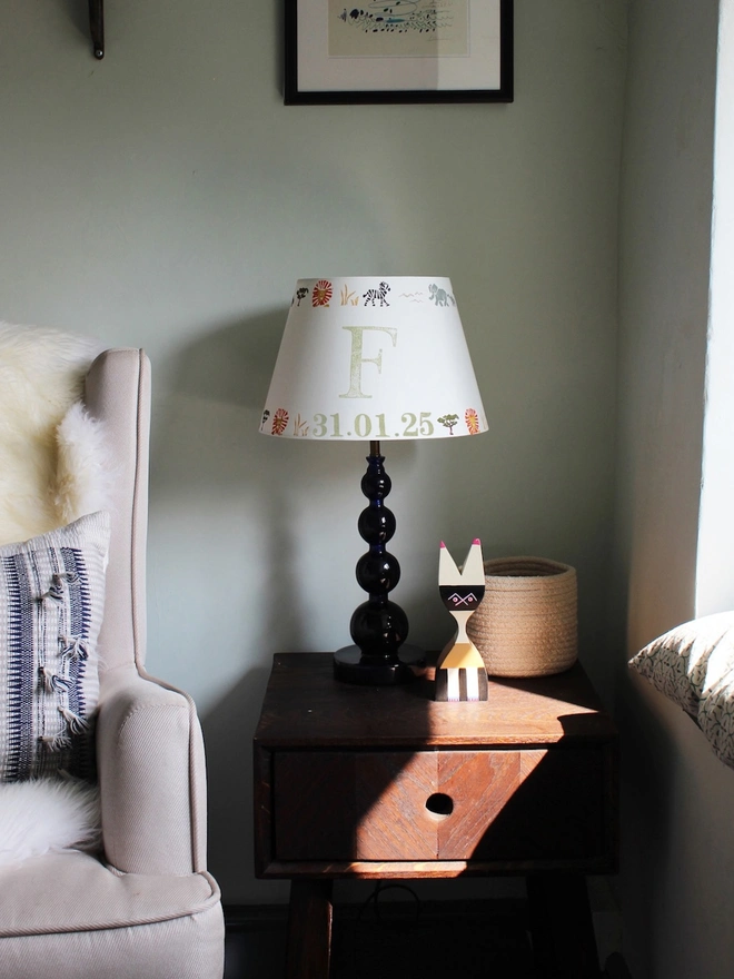 Safari lampshade with letter F, styled on side table in bright boys bedroom. Handmade safari lampshade for kids bedroom, personalised with letter F. Colourful empire light shade with safari animals, a green lampshade option for boys lampshade or kids bedroom design. The lamp is on a dark blue lamp base next to a cream arm chair with blue cushion and sheep skin. 
