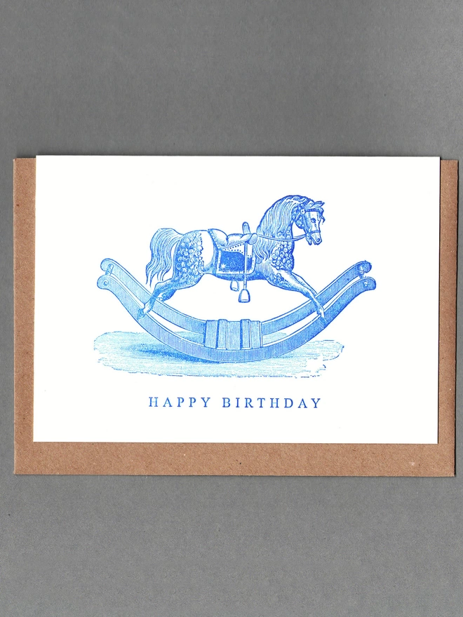 White card with blue illustration of a rocking horse and text reading 'Happy Birthday' with a brown envelope behind