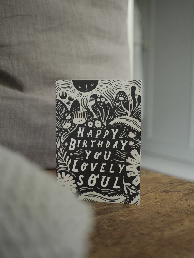Black and white greeting card with illustration and the words Happy Birthday you lovely soul written on it stood up on wooden surface with grey cushions to the side
