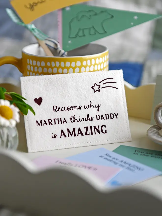 A close-up of a handmade felt envelope with the words "Reasons why MARTHA thinks DADDY is AMAZING" displayed on a white breakfast tray. Around it are pastel-coloured prompt cards with handwritten messages, a small vase of daisies, a yellow mug with crafting items, and a miniature candle on a ceramic plate, creating a cosy Father's Day setting.