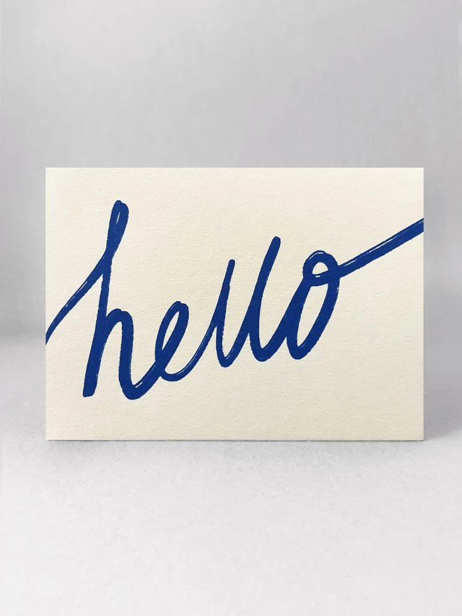 Handwritten blue hello is screenprinted on this landscape off-white greetings card. Stood front on, in a light grey studio set.