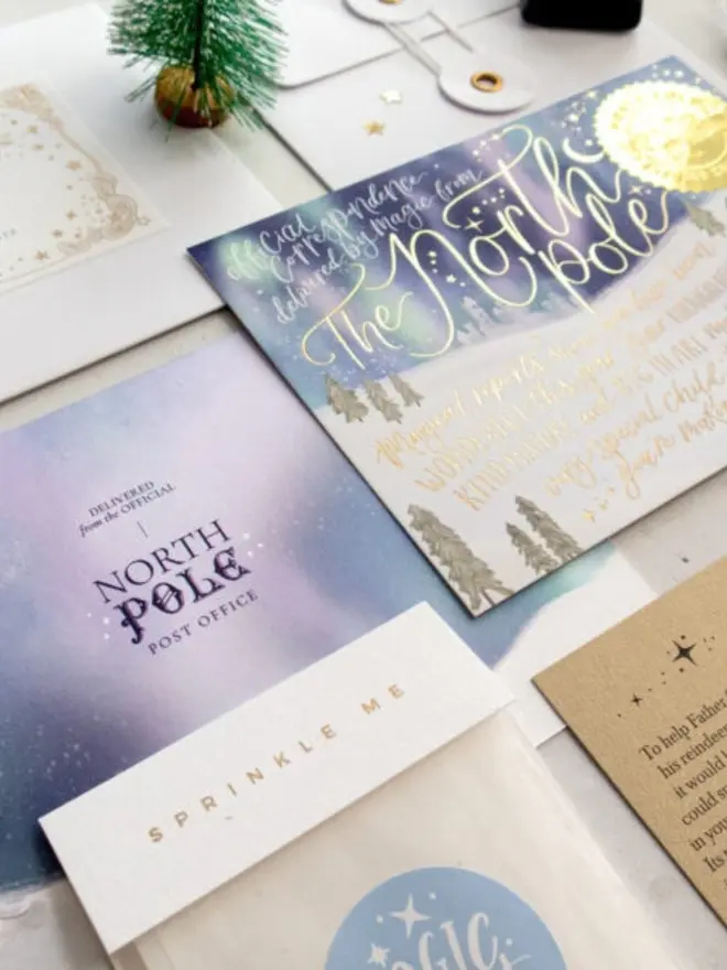 An angled view of the lilac and blue "Delivered from the official North Pole Post Office" card back, the "Sprinkle me" Magic Dust bag and the gold foil North Pole notecard, with a white envelope and a miniature frosted tree behind.