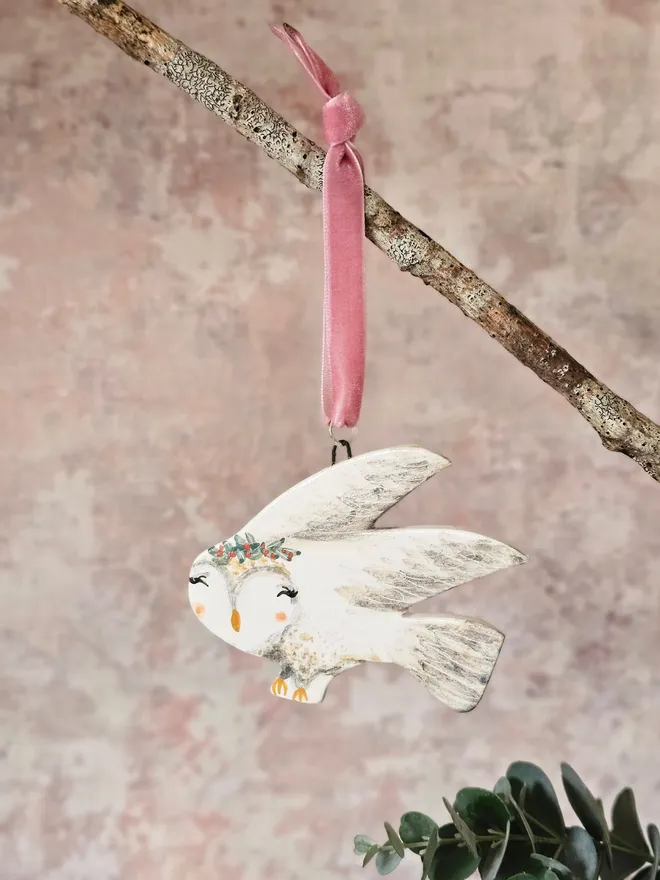 Decorative owl ornament with floral crown hanging from a branch on a soft pink background.