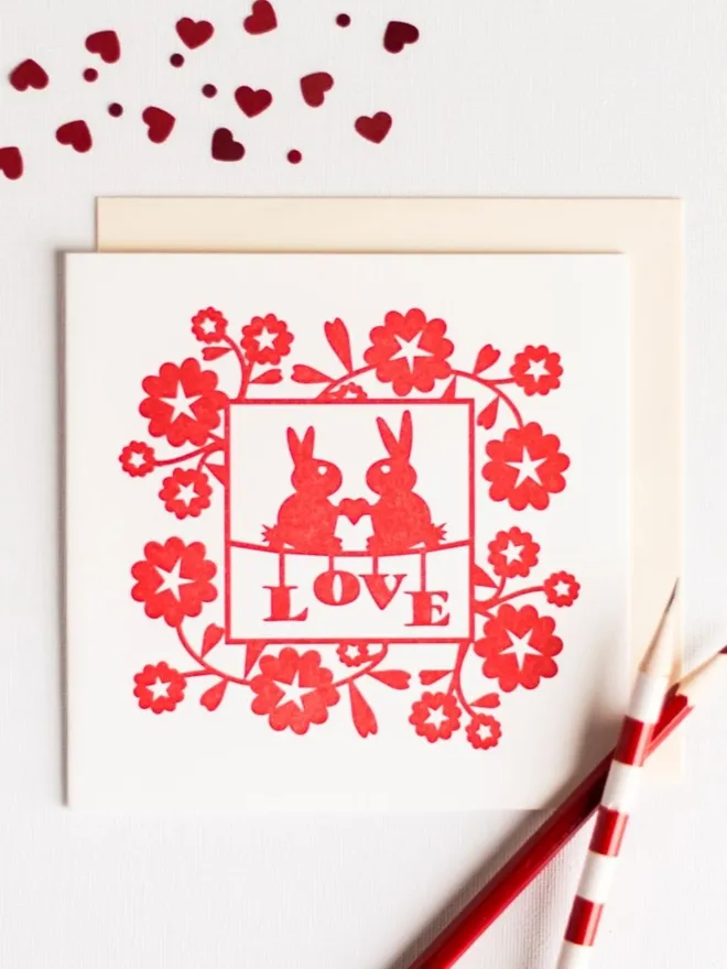 Square white card with red love bunnies design laid flat on table styled with red heart confetti and red pencils