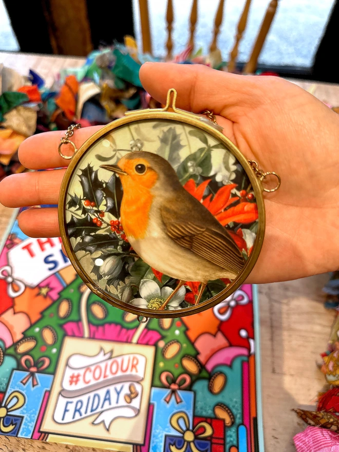 A photo showing a circular brass frame with a print of a robin amongst Christmas foliage. The frame is held in the palm of a hand to show the size and scale.