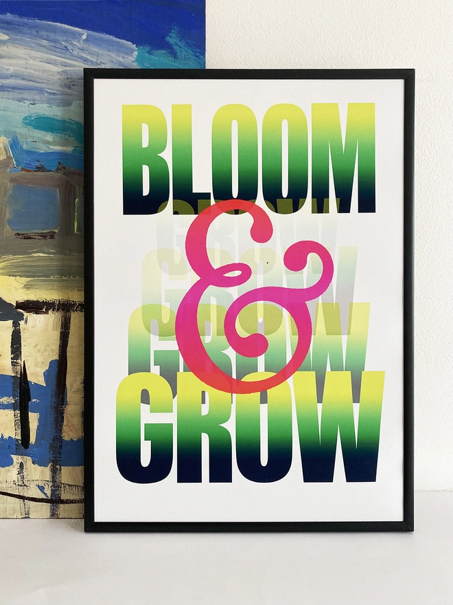 Framed multicoloured typographic print of “Bloom & Grow”  The print rests against a blue and yellow abstract painting.