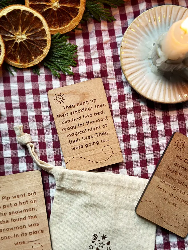 Close-up of engraved wooden Christmas story prompt cards on a red gingham cloth beside a pink book, showing a mix of story beginnings and endings for children’s festive storytelling.