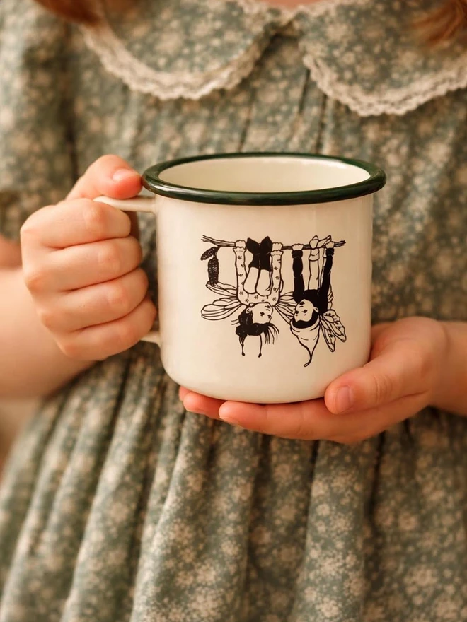 a vintage style enamel mug being held in the hands of a little girl vintage in style featuring two cute pixie pals hanging upside down on a branch