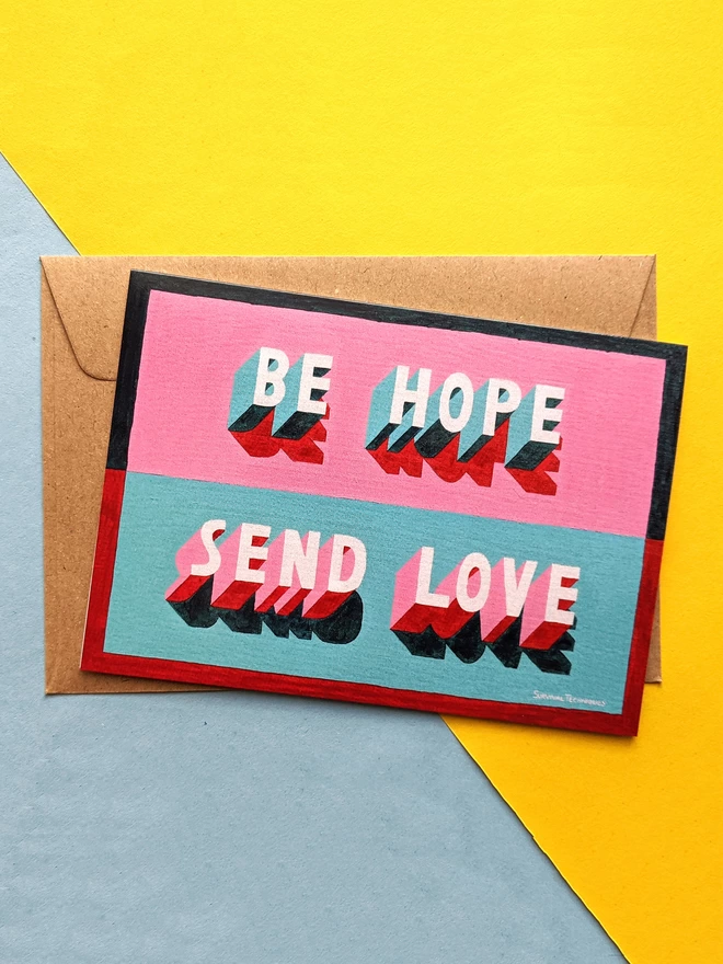 Greetings card reading Be Hope Send Love by artist Survival Techniques lies on top of a kraft envelope and a yellow and blue background