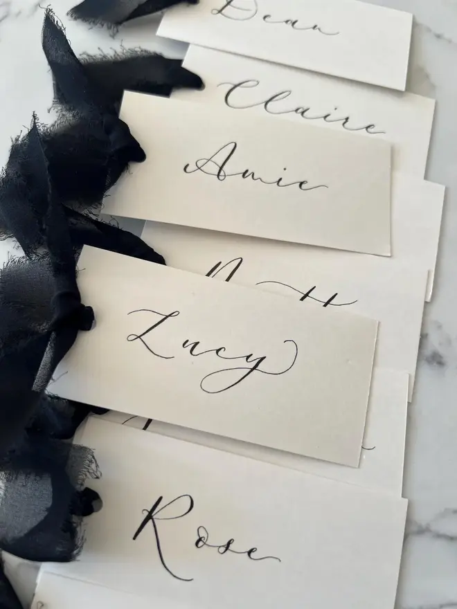 Personalised Calligraphy Wedding Cream Place Cards, cream place cards set together on a marble surface with black ribbons 