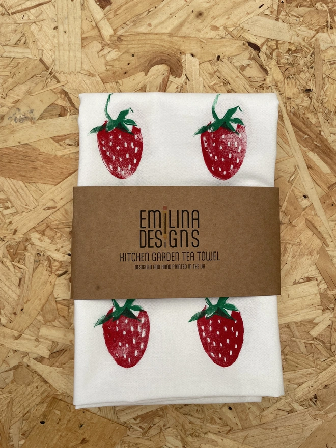 Strawberry print kitchen tea towel folded on a wooden chip table with a cardboard packaging band.