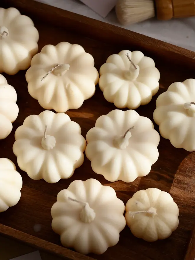 Medium Natural Wax Pumpkin Candle. A wooden tray filled with cream pumpkin-shaped candles in a mix of sizes, each with a moulded stalk and cotton wick. The elegant sculptural candles make a beautiful neutral centrepiece for autumn tables and Thanksgiving décor.