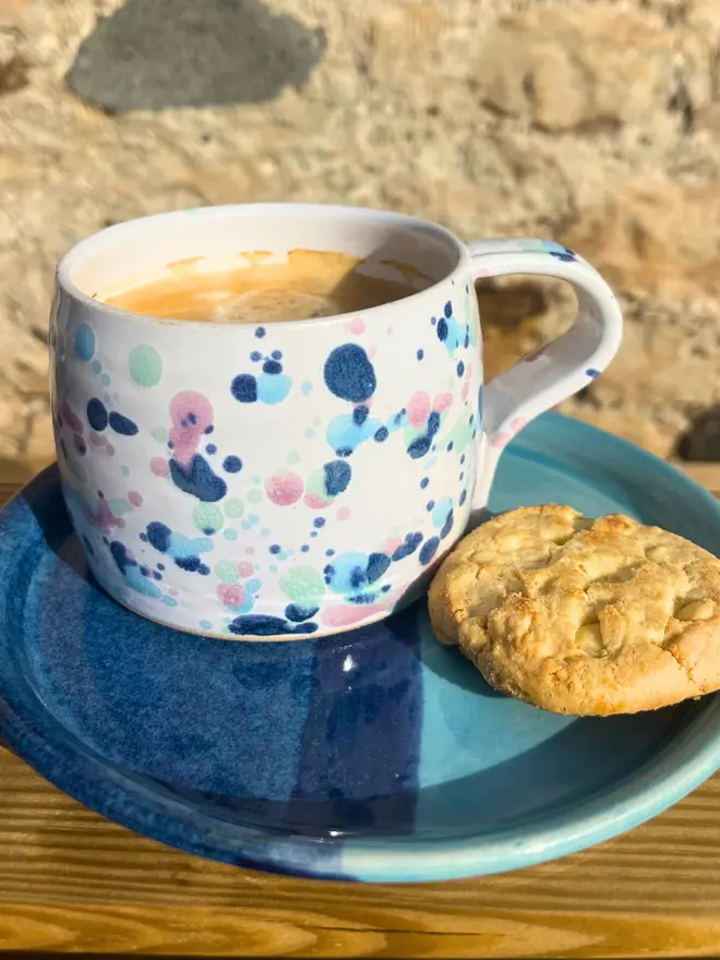 Sweetpea Large Round Mug, a colourful mug sitting on a wooden surface against a brick backdrop. It is accompanied by a plate and some biscuits. 