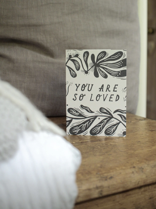 Black and white greeting card with illustration and the words you are so loved on it lay on top of a wooden surface