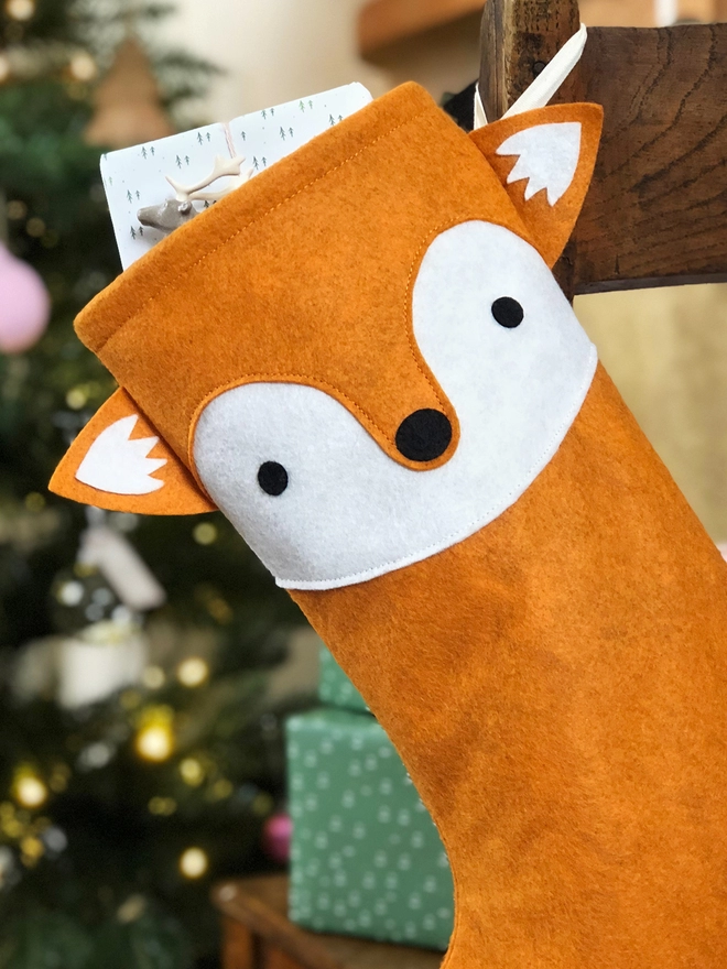 A handmade felt fox stocking hangs on a wooden chair, where a little girl sits, in front of a Christmas Tree.