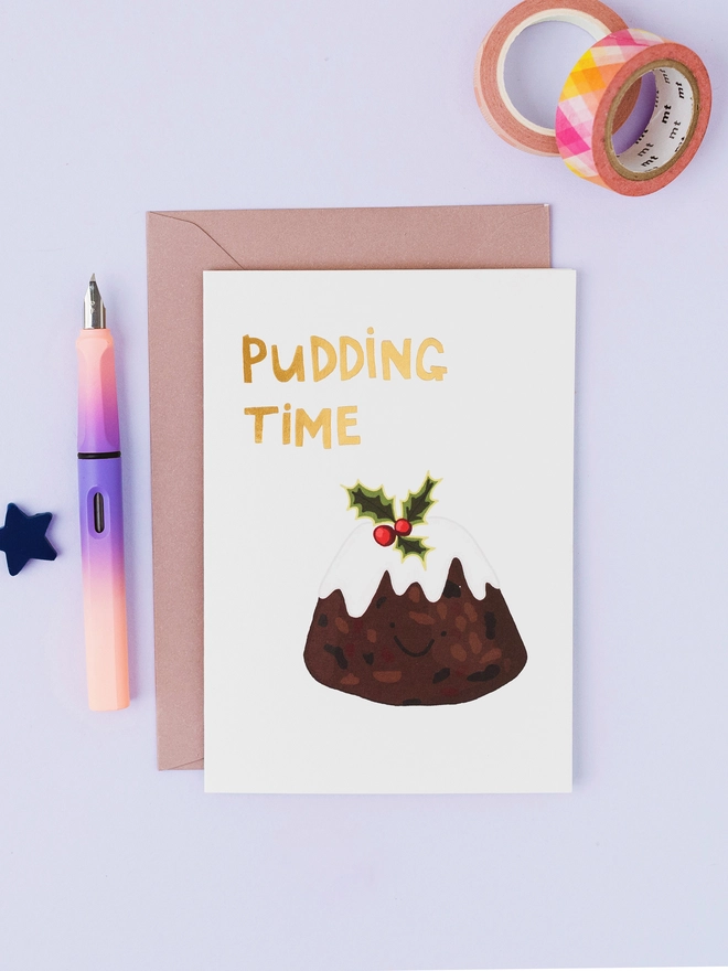 White Christmas greeting card featuring a hand-drawn Christmas pudding with white icing, holly and gold foil Pudding Time lettering.