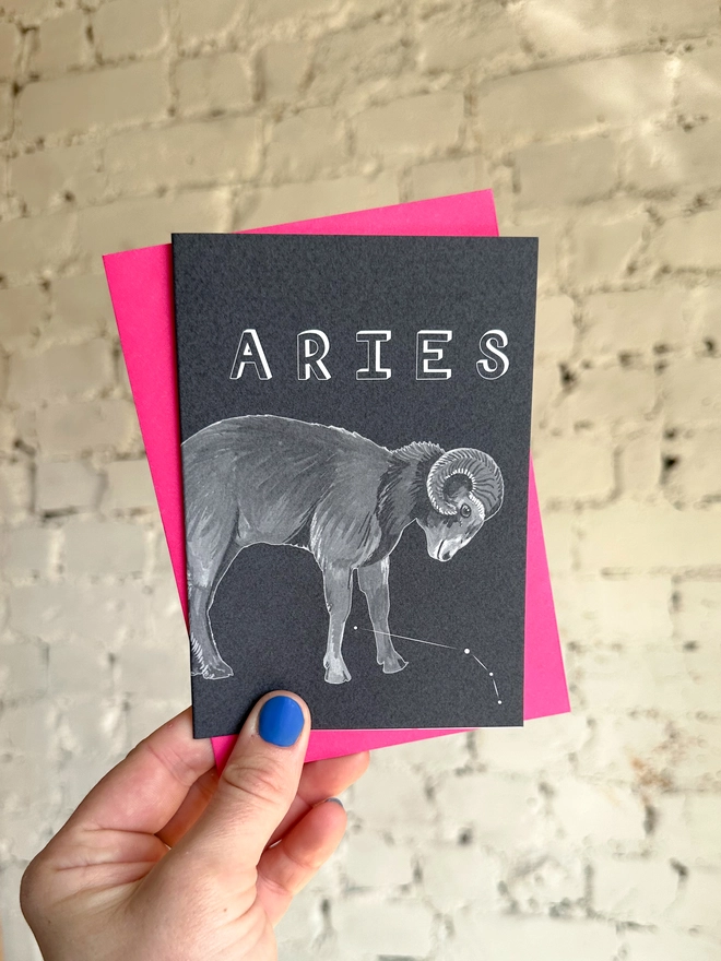 a greetings card featuring a greyscale illustration of a zodiac style ram with the text “ARIES” with a neon pink envelope